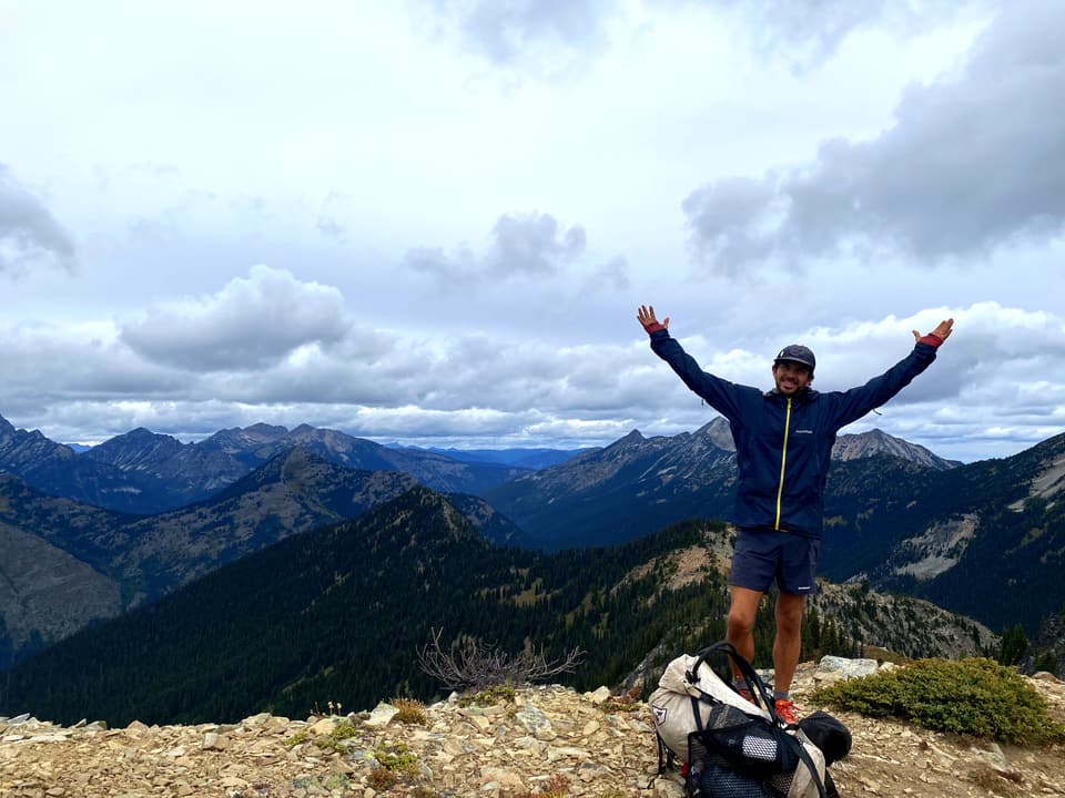 Atlas with his arms up on a rocky summit above the last North Cascades ridges, day 129